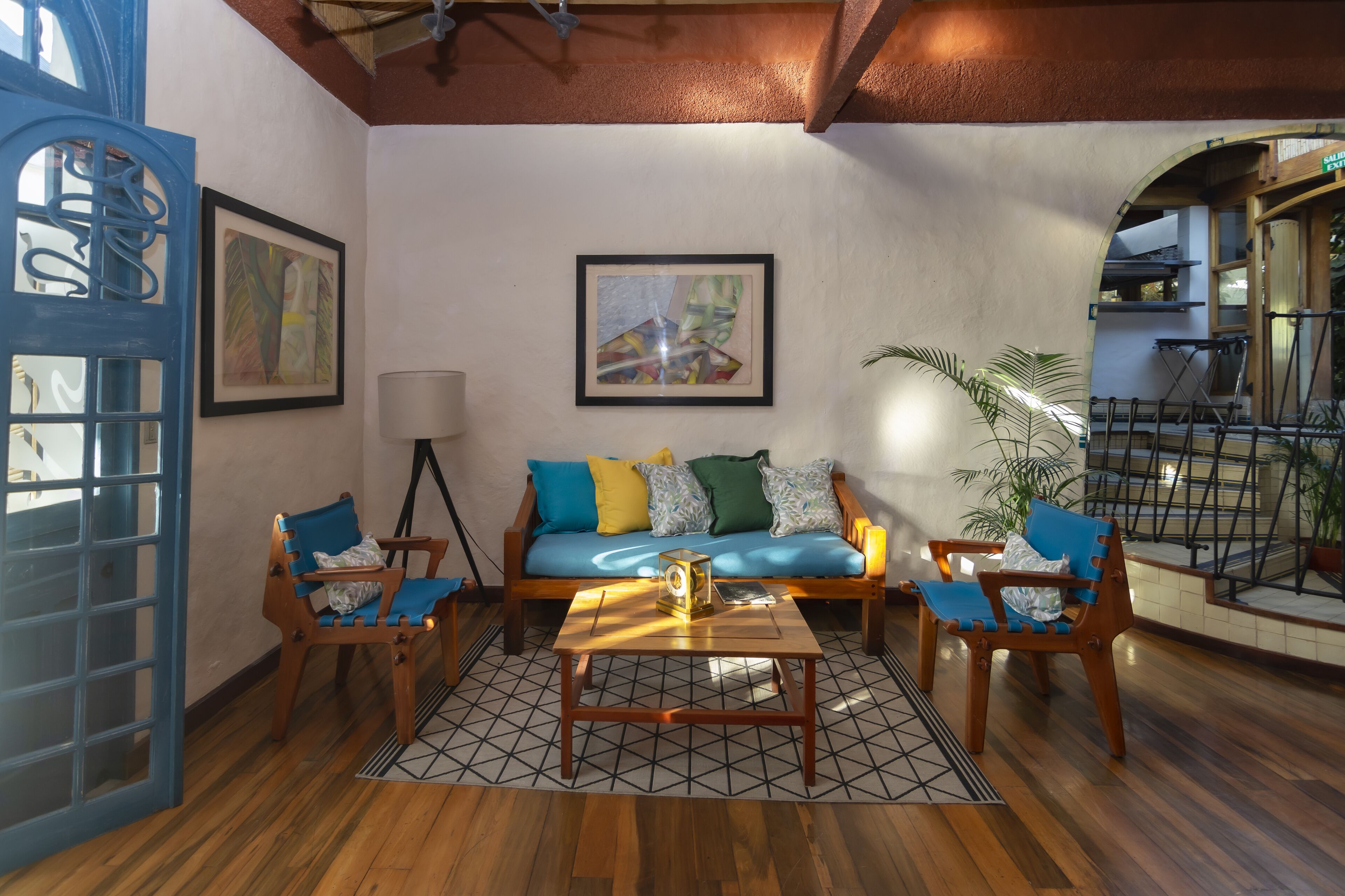 A cozy living room with a wooden floor, a blue sofa with colorful cushions, two wooden chairs, a coffee table, and a framed abstract painting on the wall.