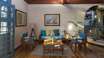 A cozy living room with a wooden floor, a blue sofa with colorful cushions, two wooden chairs, a coffee table, and a framed abstract painting on the wall.