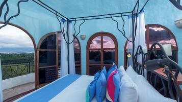 Un dormitorio con cama, almohadas, una mesa y sillas. Hay dos ventanas con cortinas, y se aprecia la vista del paisaje a través de ellas.