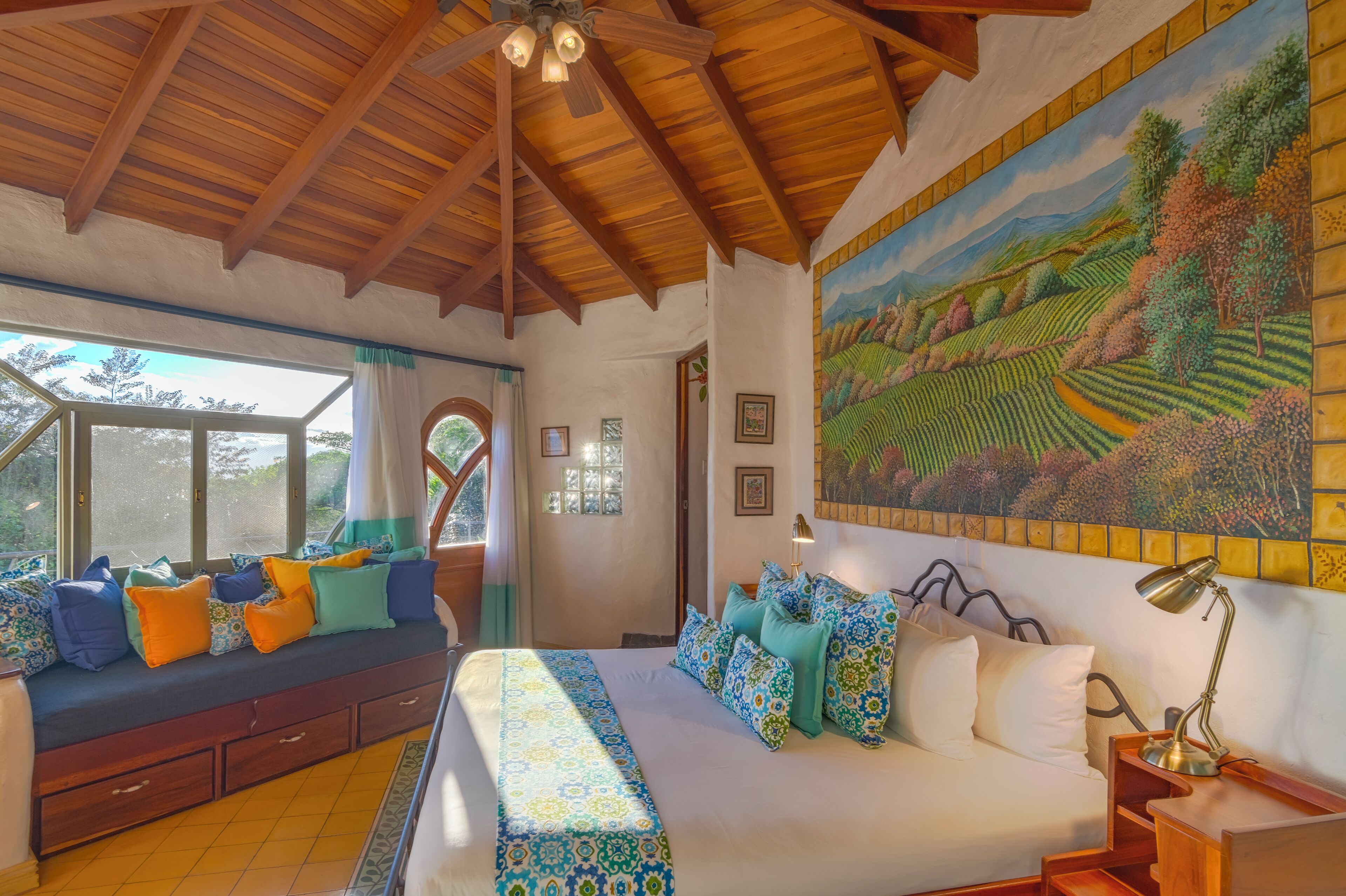 A bedroom with a large bed, a wooden ceiling, a mural, and a view through a window.