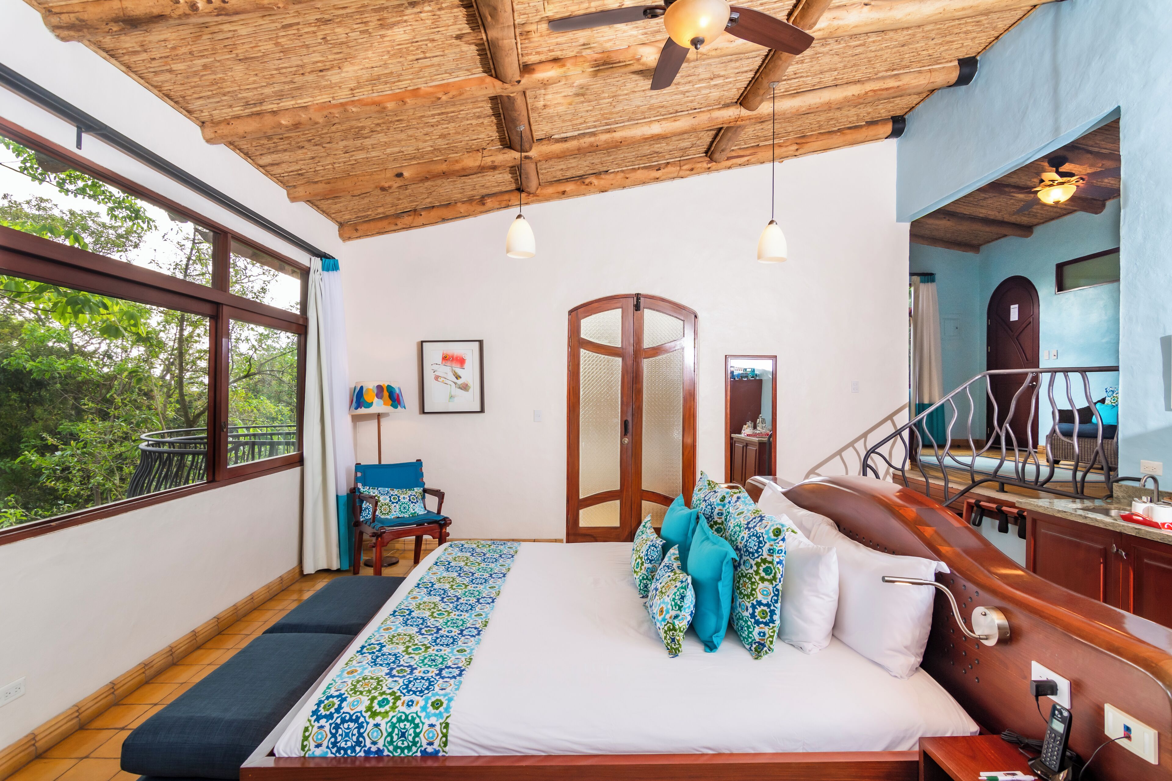 A bedroom with a large bed, wooden ceiling, and a view of trees through a window.