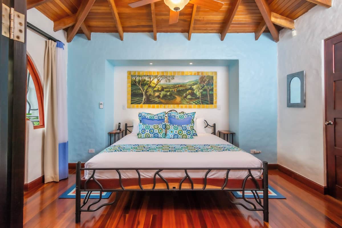 A bedroom with a bed, wooden floor, a ceiling fan, a painting, and a door.