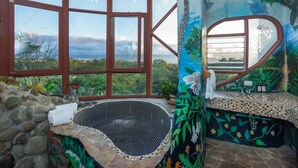 A bathroom with a large window, a stone wall, a bathtub, and a sink with a tiled countertop.
