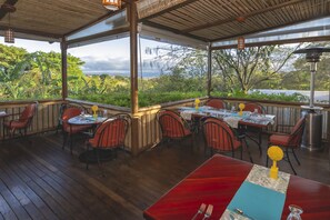 An outdoor dining area with wooden tables and chairs, surrounded by lush greenery and a view of the landscape.