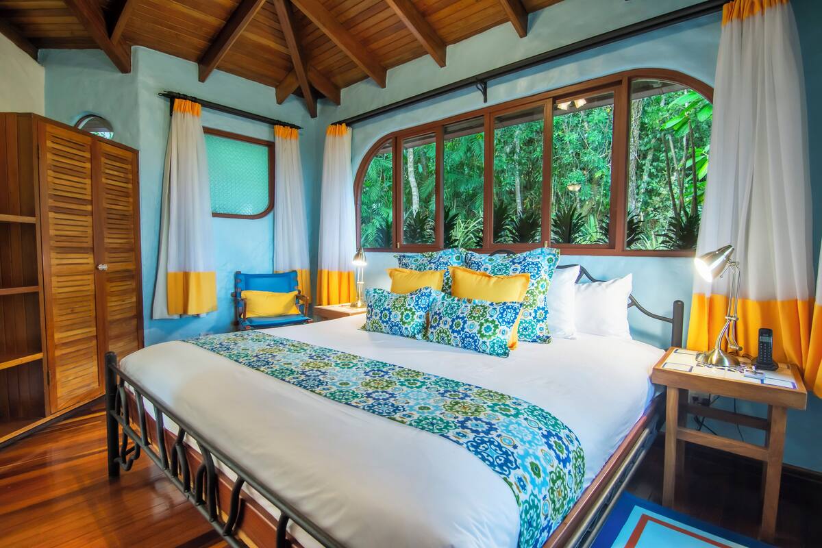 A bedroom with a large bed, wooden wardrobe, and a window with a view of greenery.