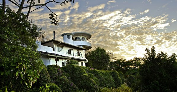 A large, white, multi-story house with a unique, rounded tower-like structure, surrounded by lush greenery and trees.