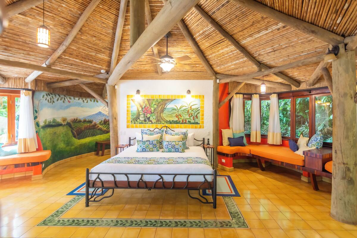 A room with a bed, a mural, a window with curtains, and a wooden ceiling.