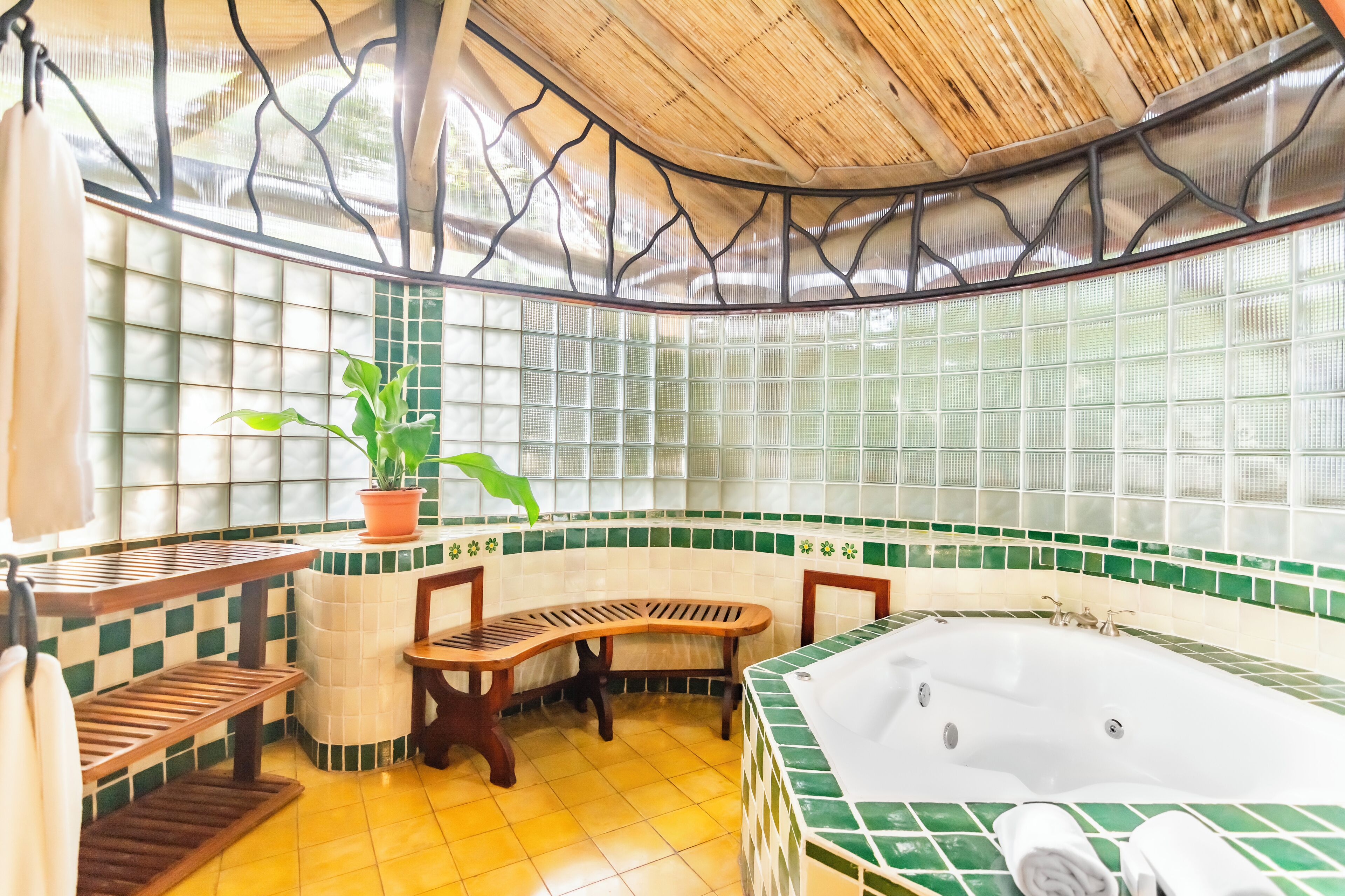 A bathroom with a bathtub, wooden benches, a potted plant, and a tiled wall with a green border.