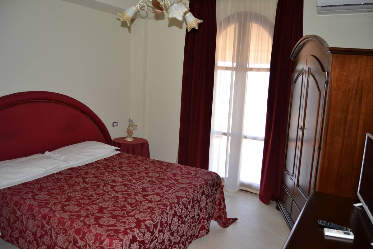 A bedroom with a large bed, a wooden wardrobe, a small table with a lamp, and a television on a wooden dresser.