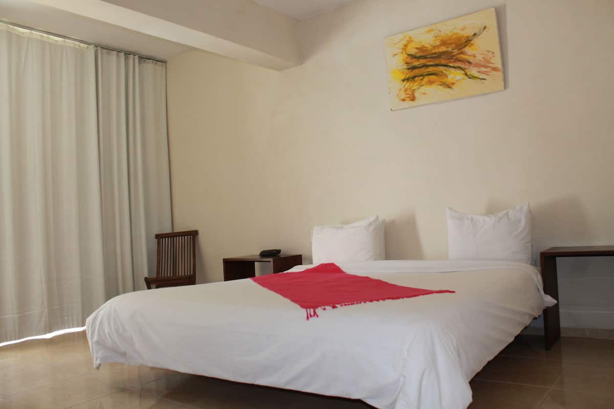A hotel room with a large bed, a red throw blanket, a wooden chair, a nightstand, and a painting on the wall.