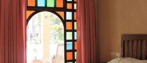 A hotel room with a bed, a ceiling fan, and a view through a window with colorful window shutters.