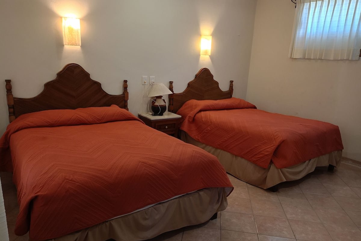 A hotel room with two beds, each with a wooden headboard and orange bedding. There are wall-mounted lights and a window with blinds.