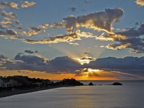 A coastal sunset with clouds, calm sea, and buildings along the shore.