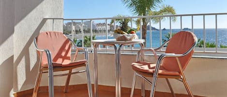 A balcony with two metal-framed chairs and a small table, overlooking a coastal view with palm trees and buildings.