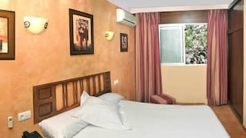 Una habitación de hotel con cama, teléfono, lámparas fijadas a la pared y ventana con cortinas.