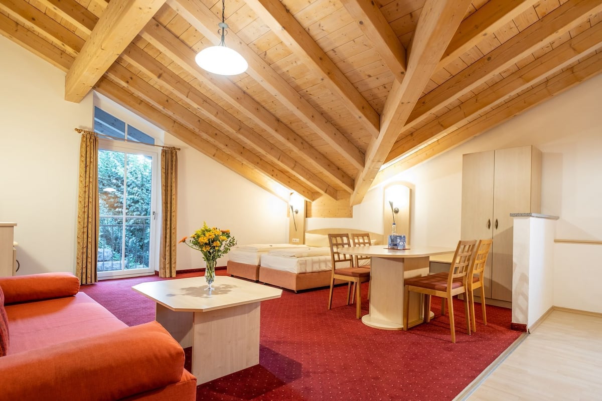 A cozy room with a wooden ceiling, a bed, a dining table with chairs, a small table with a vase, and a red carpet.