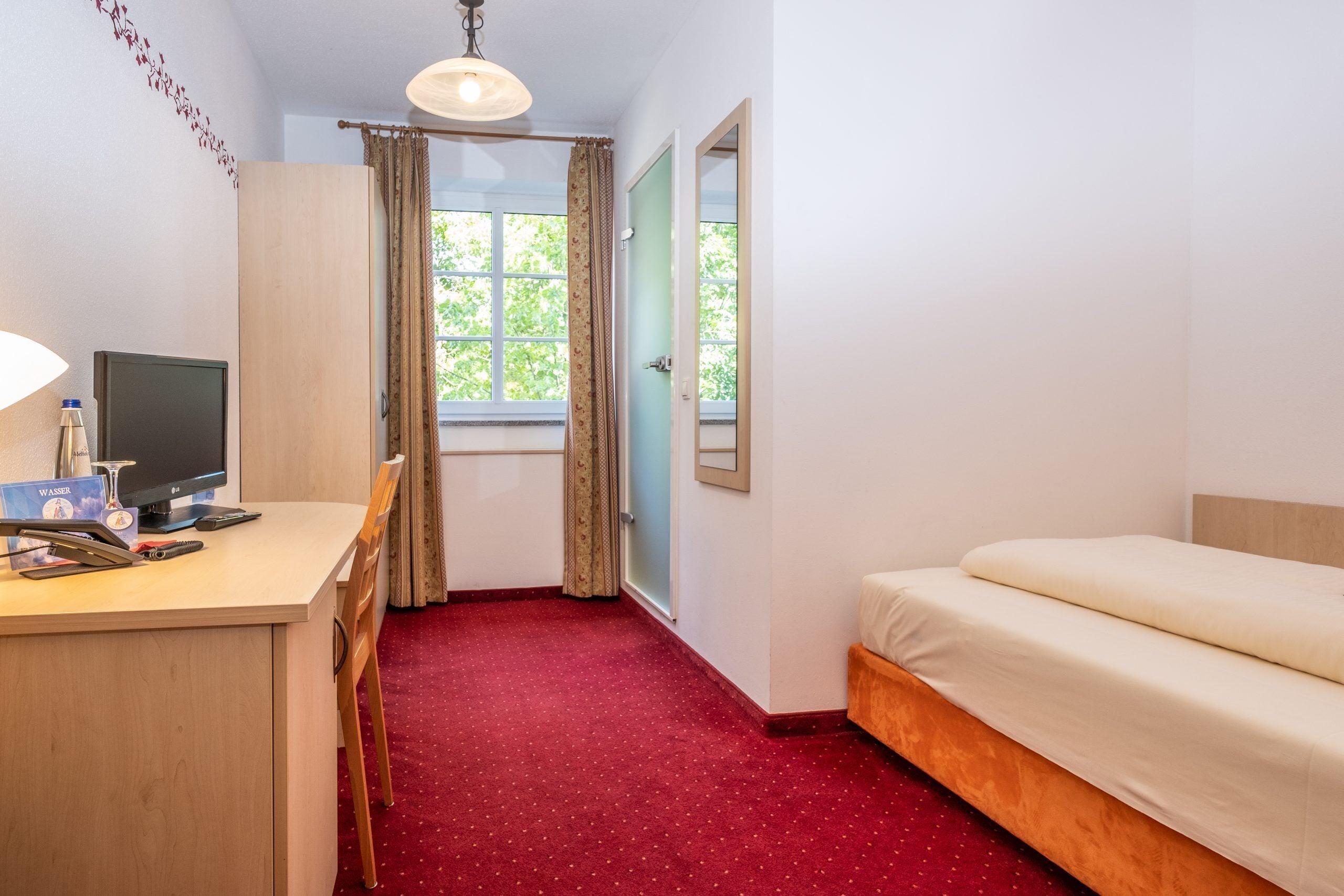 Una habitación de hotel con escritorio, cama, ventana con cortinas y suelo cubierto de moqueta roja.