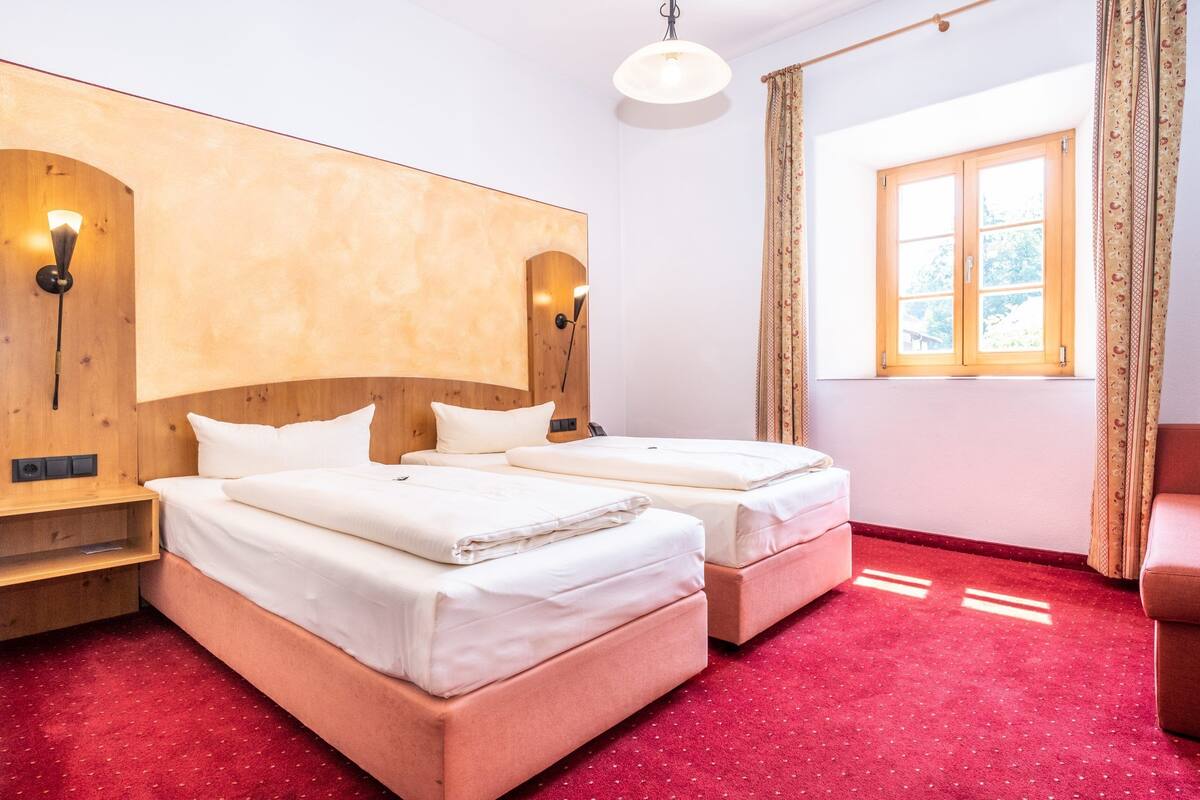 A hotel room with two beds, wooden headboards, a red carpet, a window with curtains, and wall-mounted lights.