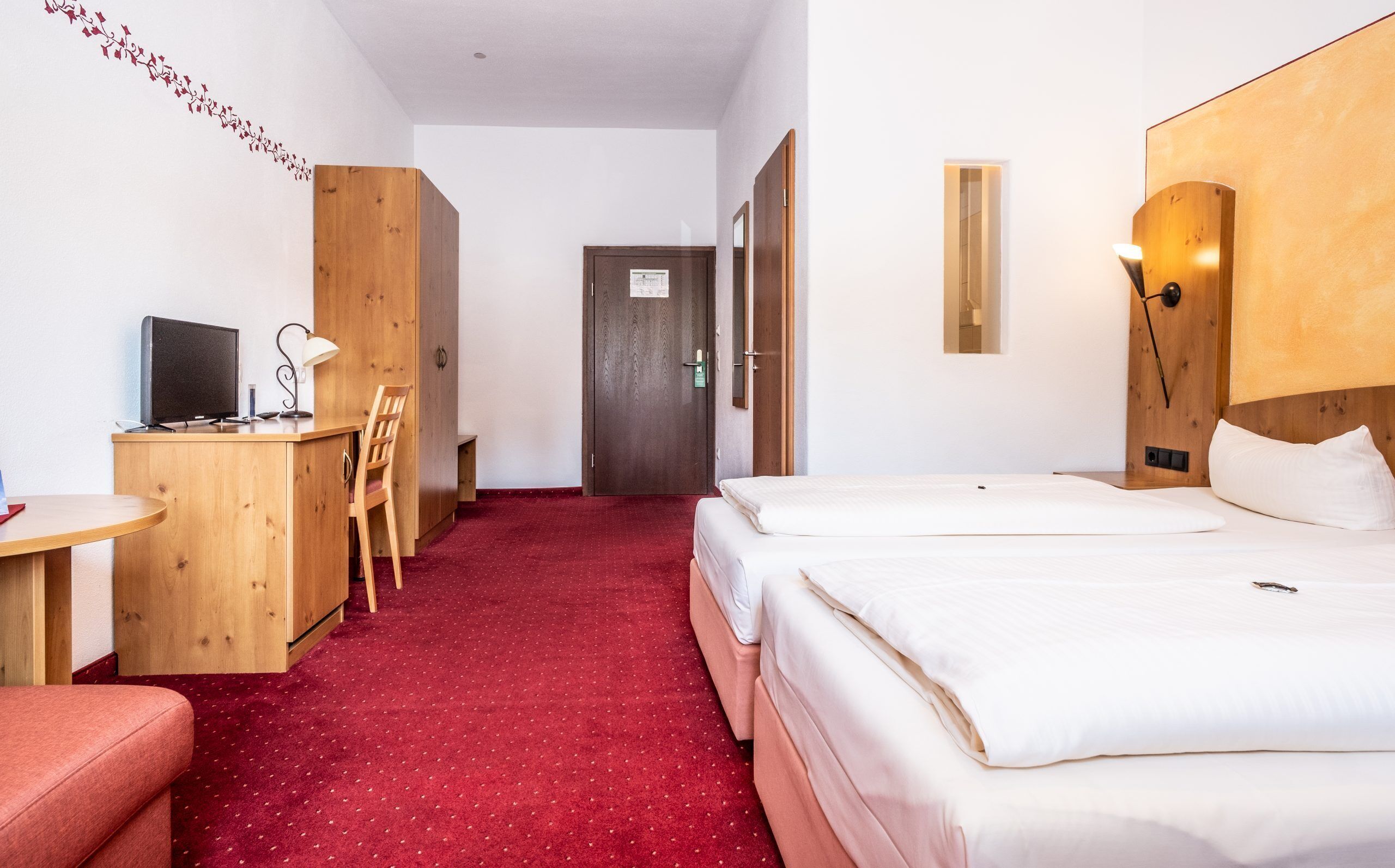 A hotel room with a desk, chair, two beds, a red carpet, and a wooden wardrobe.