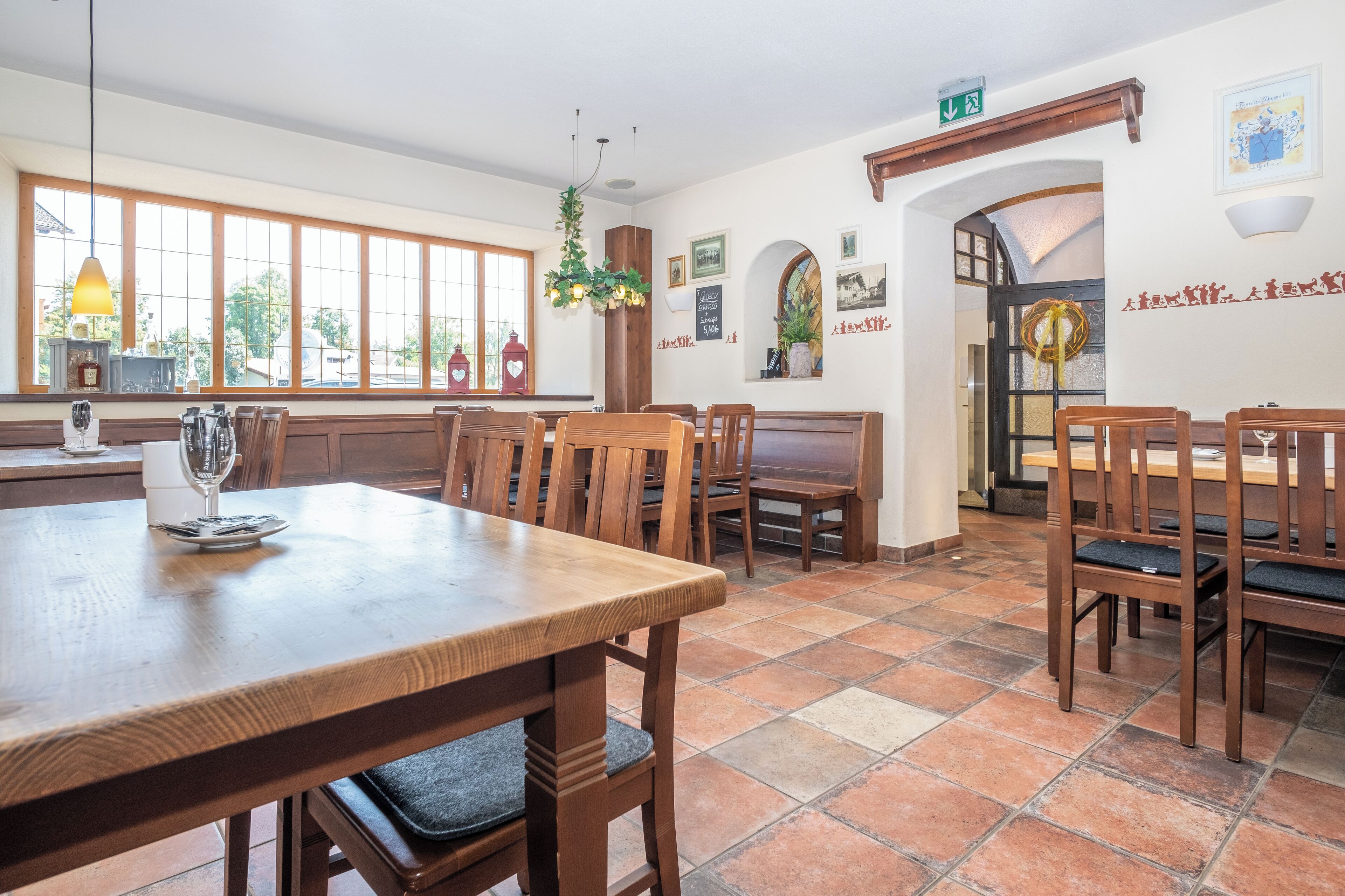 Un espace repas avec des tables et des chaises en bois, de grandes fenêtres et un sol carrelé.