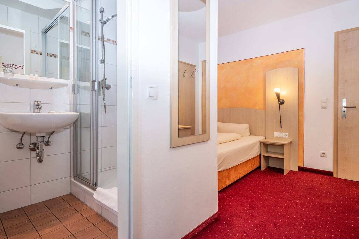 A hotel room with a shower, sink, bed, bedside table, and a mirror.
