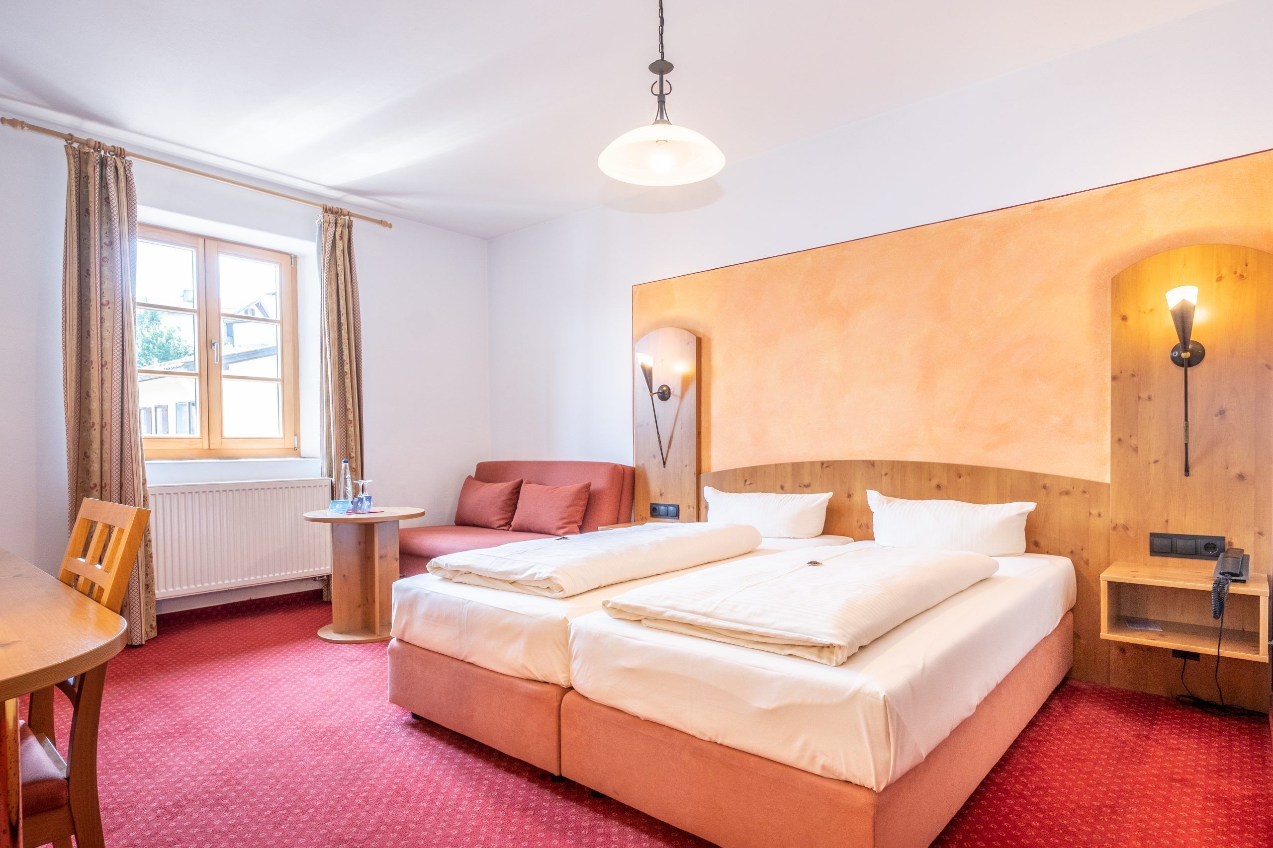 A hotel room with a large bed, a small table, a chair, a window with curtains, and a wall-mounted light fixture.