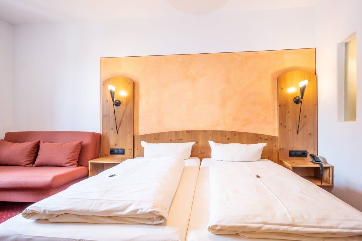 A hotel room with two beds, a red sofa, wooden headboard, and wall-mounted lamps.