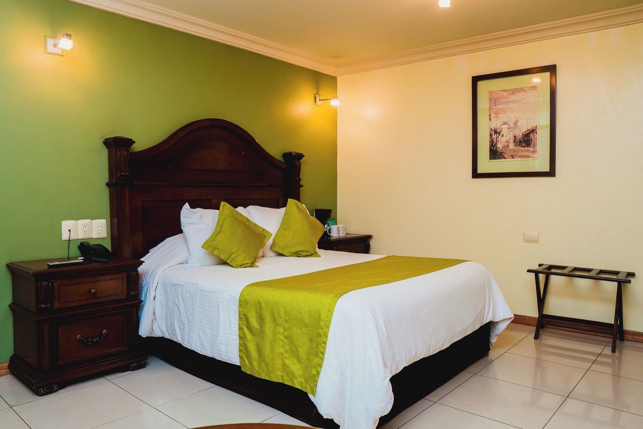 A neatly made bed with a green and white bedspread, a wooden nightstand with a phone and a lamp, and a framed picture on the wall.