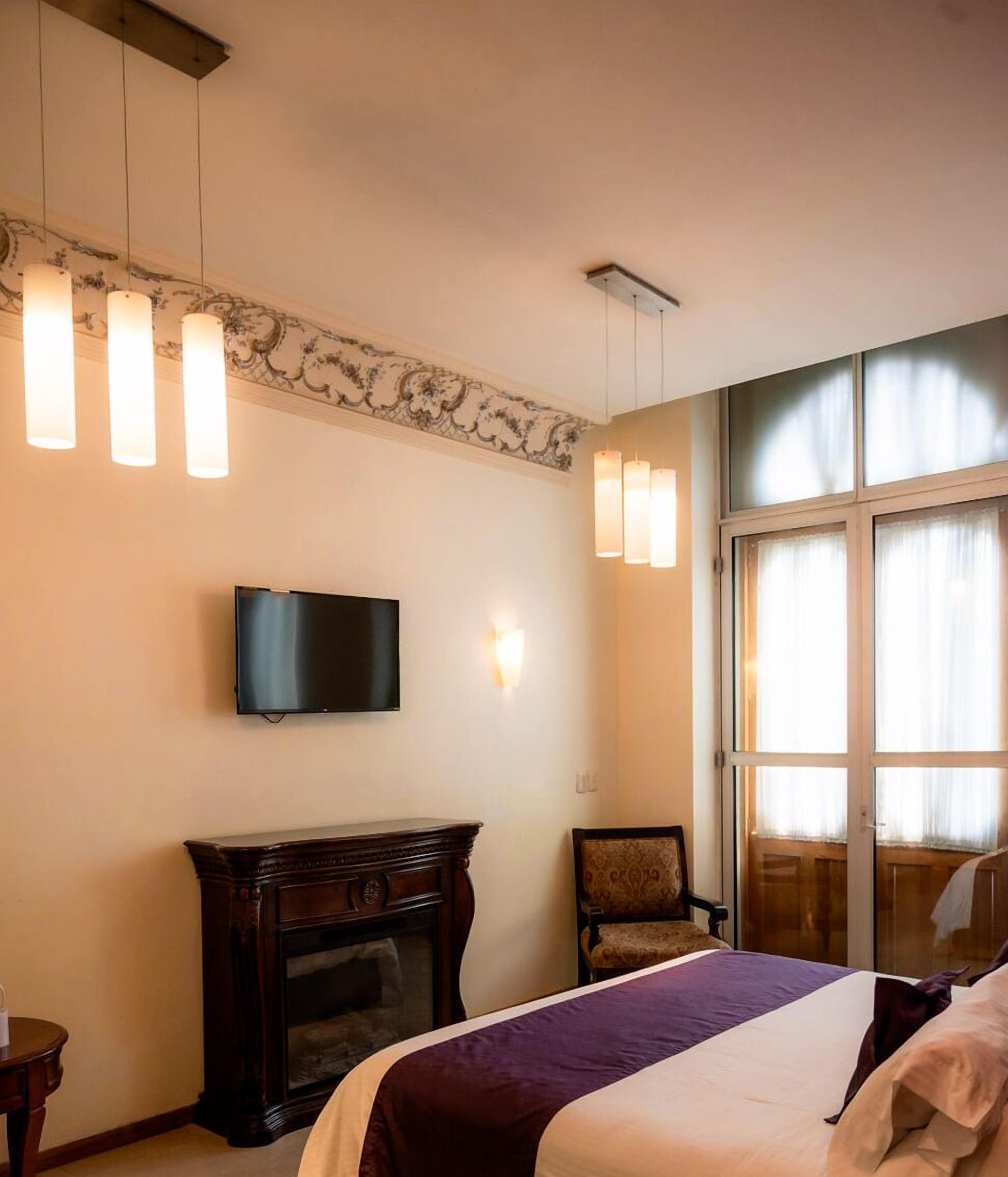 Una habitación de hotel con cama, televisor montado en la pared, chimenea y ventana con cortinas.