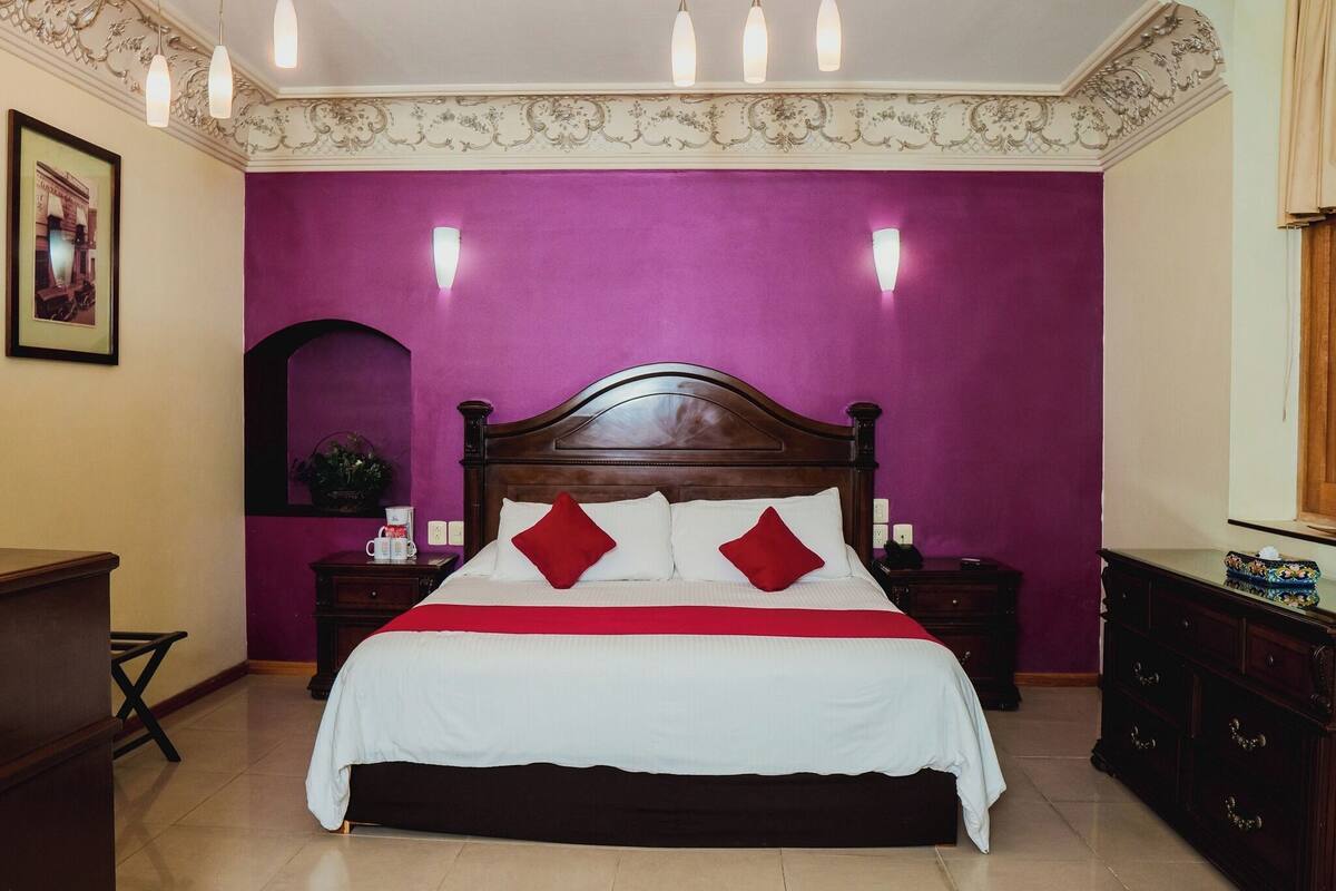 Une chambre à coucher avec un grand lit, des murs violets et une tête de lit en bois. On y trouve deux tables de chevet avec des lampes, une commode et un tableau encadré accroché au mur.