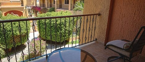 A balcony with a glass table and a chair, overlooking a courtyard with palm trees and a building with arches.
