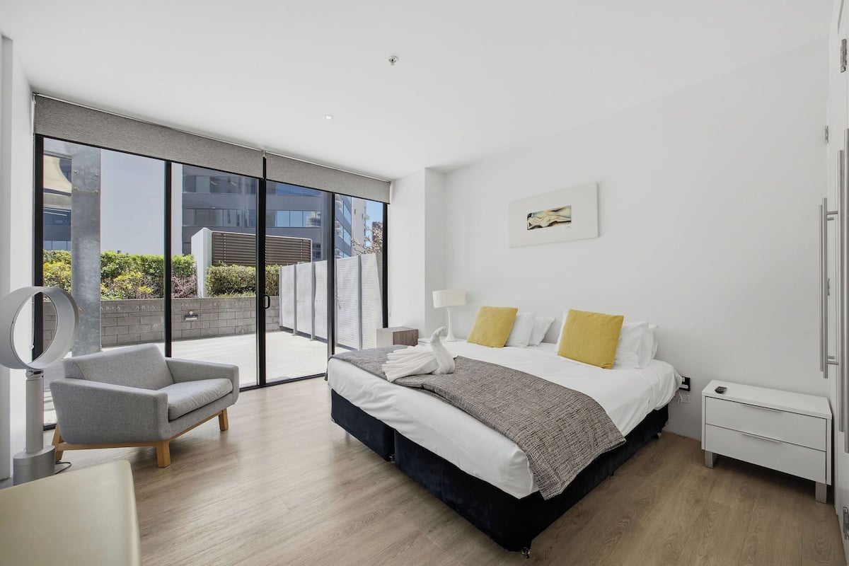 A modern hotel room with a large bed, a gray armchair, a bedside table, and a view of the outside.