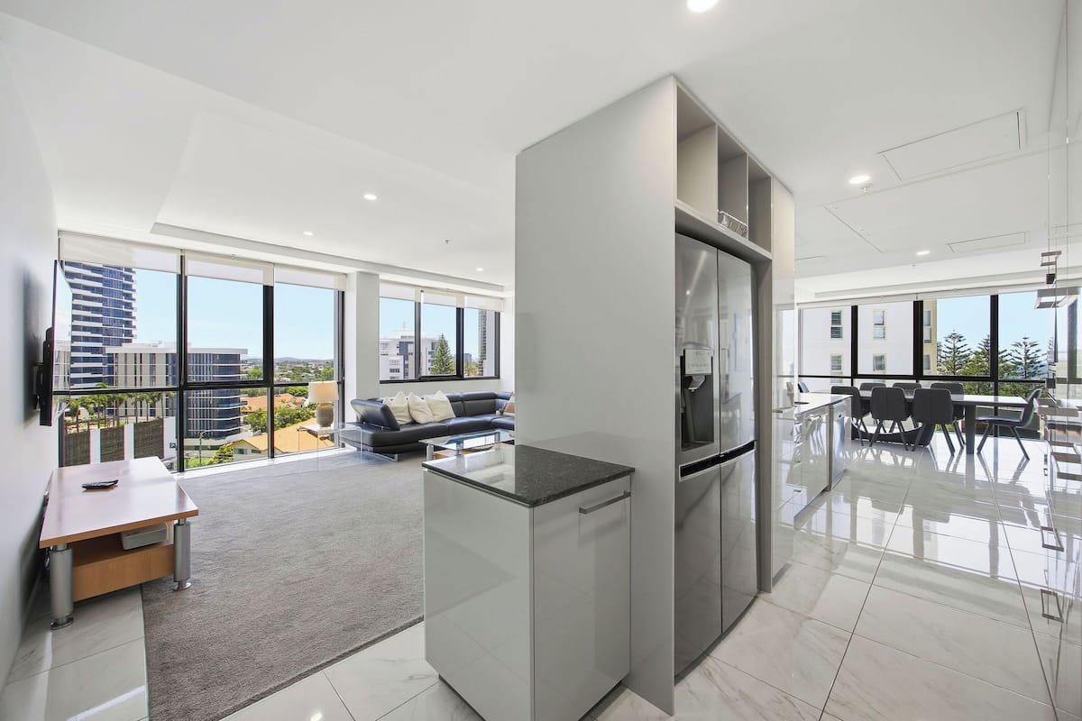A modern kitchen with a refrigerator, a dining area with chairs, and a sofa in a spacious living area with large windows.