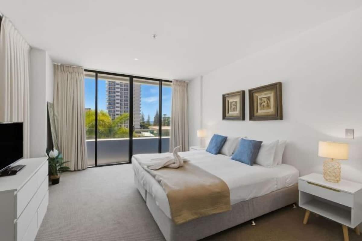 A hotel room with a bed, a TV, a desk, a lamp, and a view of a building through a window.