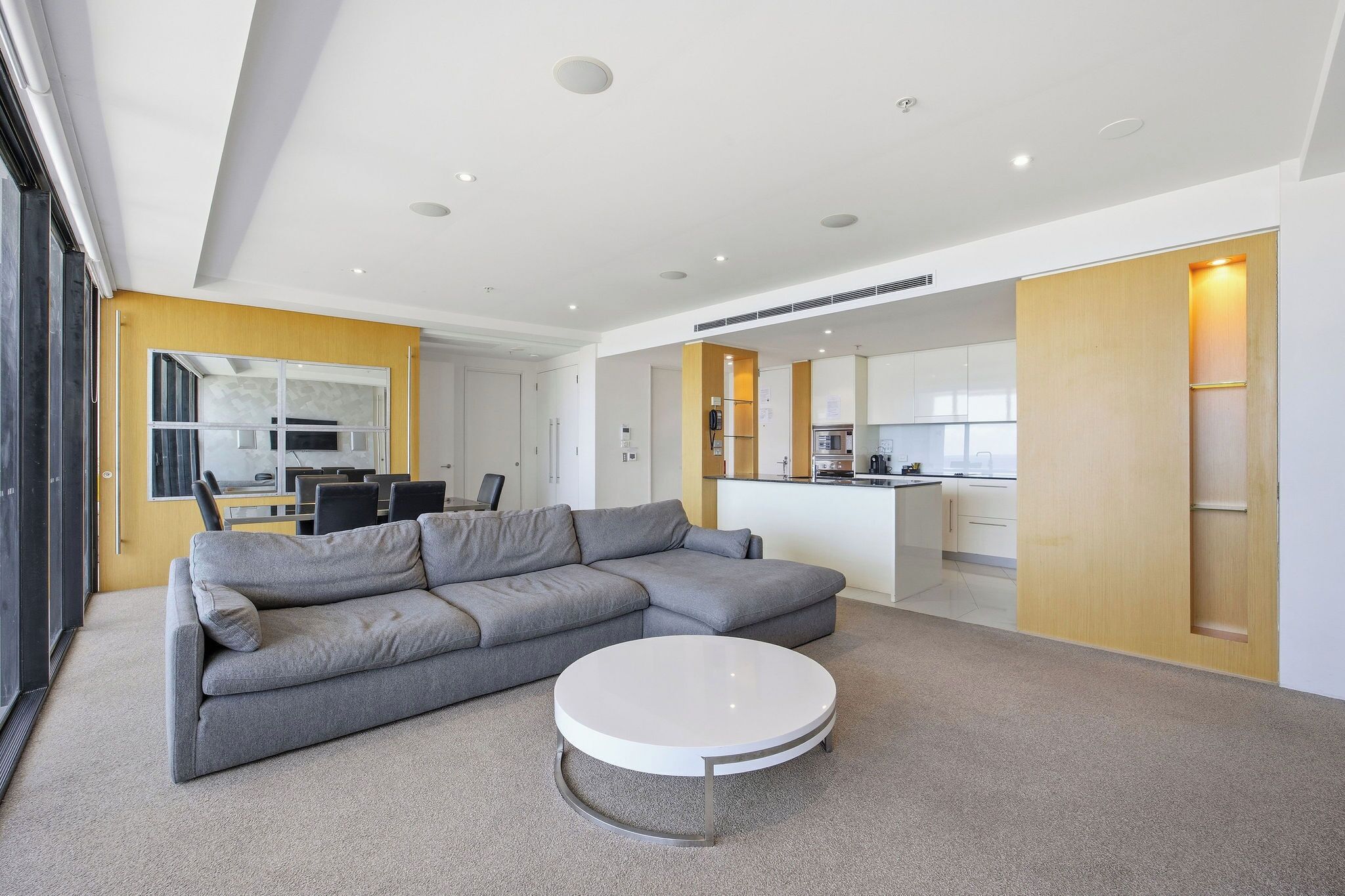 Ein modernes Wohnzimmer mit einem grauen Ecksofa, einem runden Couchtisch und einer Küche im Hintergrund.