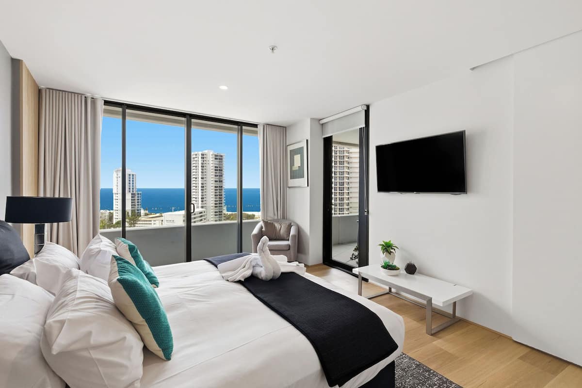 A modern hotel room with a large bed, a flat-screen TV, a bench, and a view of the city and ocean.