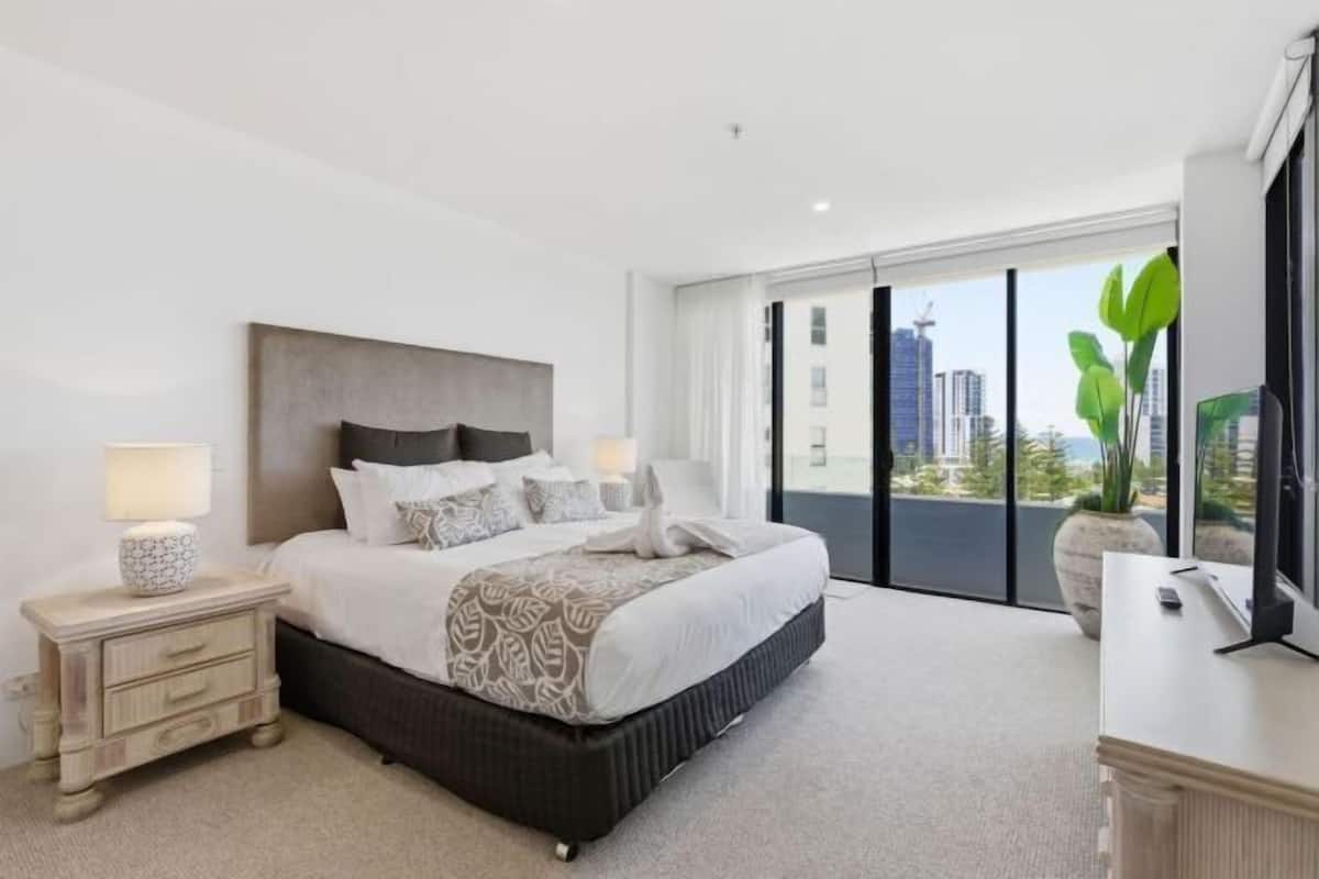 A modern hotel bedroom with a large bed, bedside tables, a nightstand, a dresser, and a view of the city.