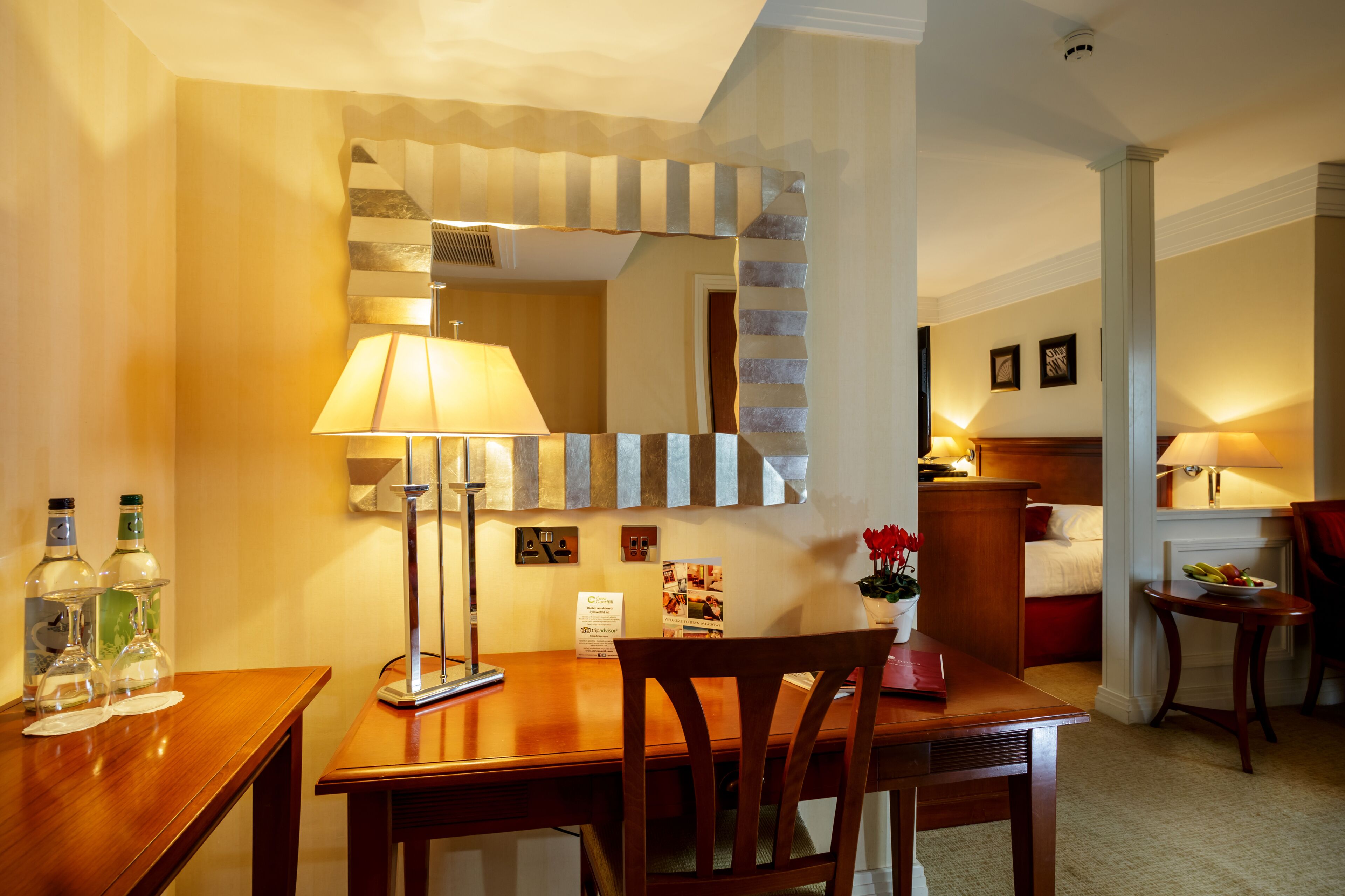 Une chambre d’hôtel avec un bureau en bois, un miroir, un lit, une table de chevet et une petite table avec une lampe.