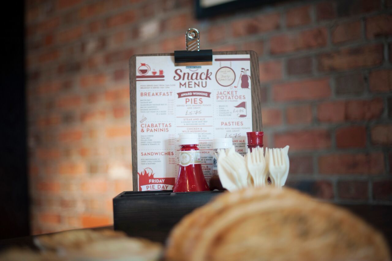 A snack menu board with items like breakfast, pies, and pasties, displayed on a clipboard against a brick wall.