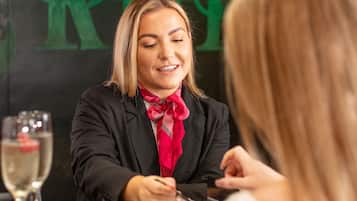 Una persona con un blazer negro y una bufanda roja sentada a una mesa con una copa de champán, frente a un muro verde con un logotipo.