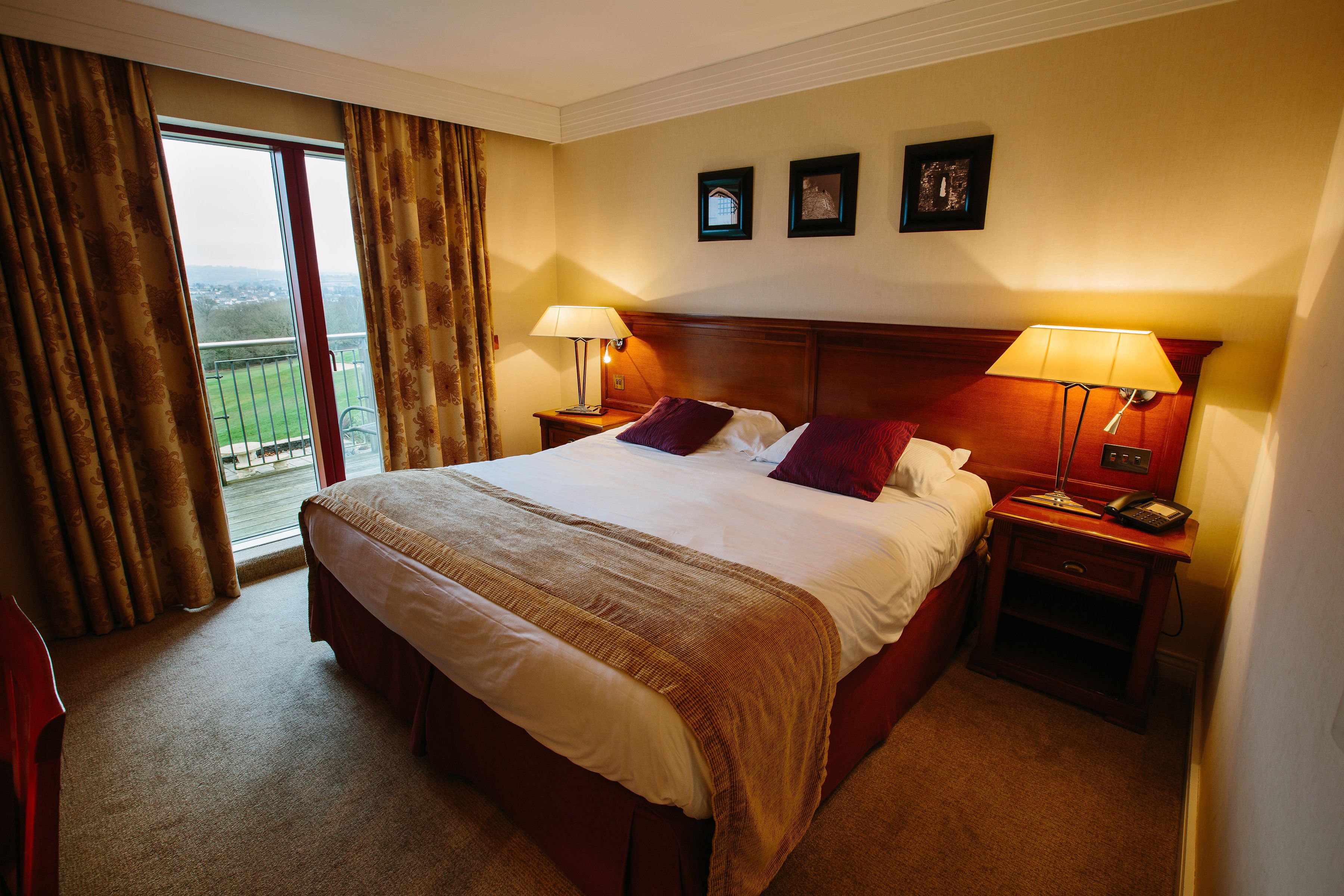 A hotel room with a large bed, two bedside tables with lamps, a view of the outdoors, and two framed pictures on the wall.