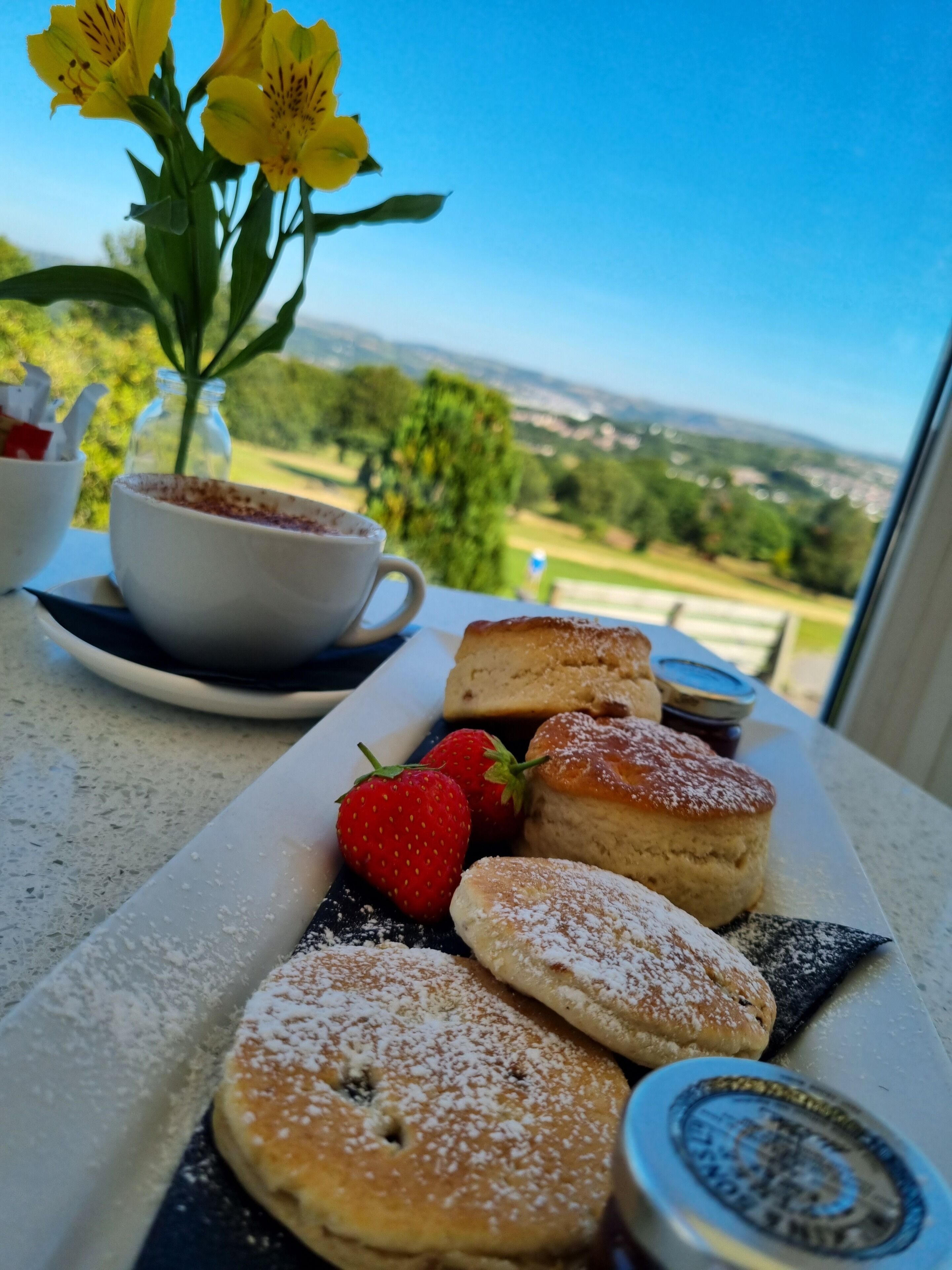 A table with pastries, strawberries, and a cup of coffee with a view of greenery and buildings.