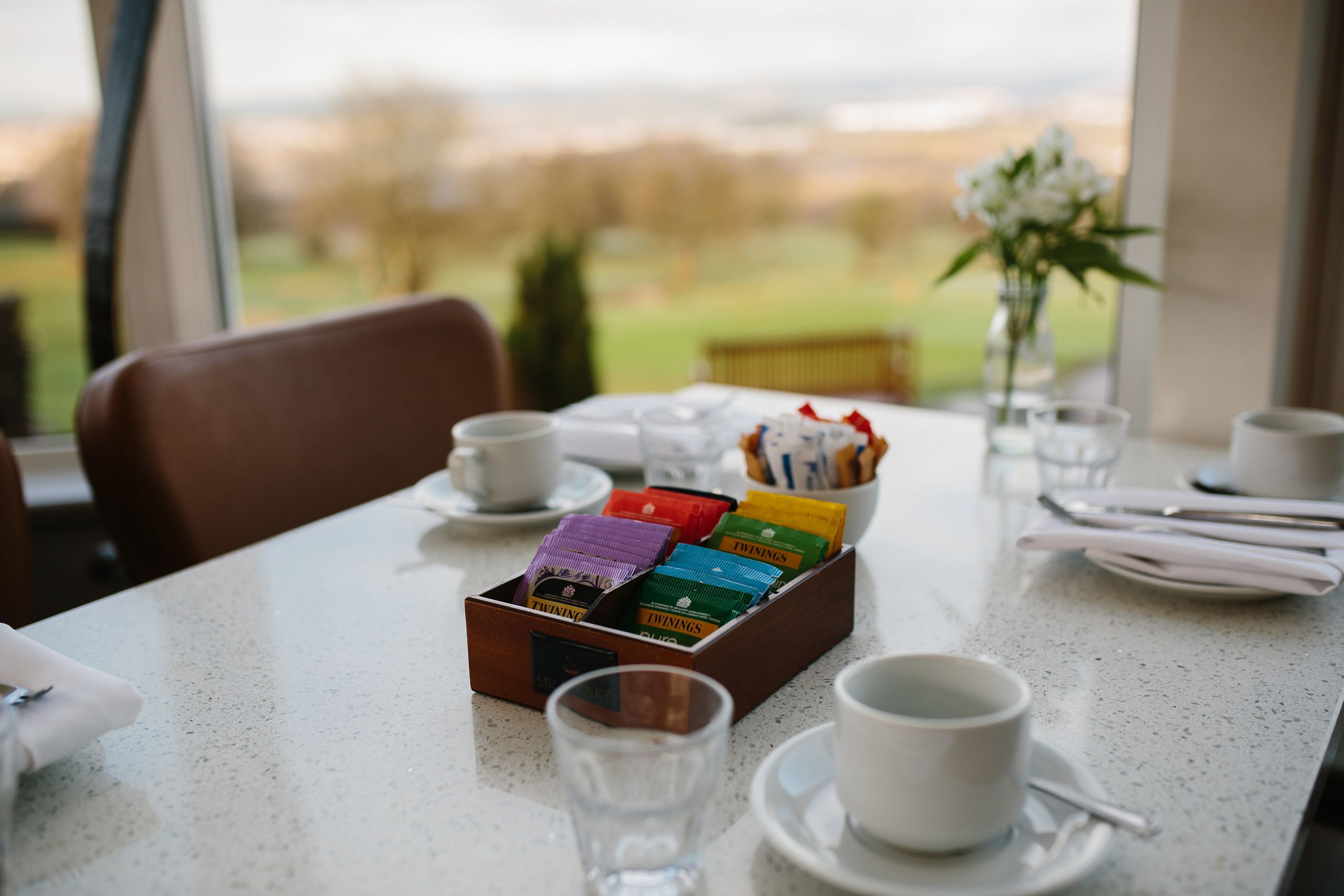 A dining table with a box of tea bags, cups, and a vase with flowers.