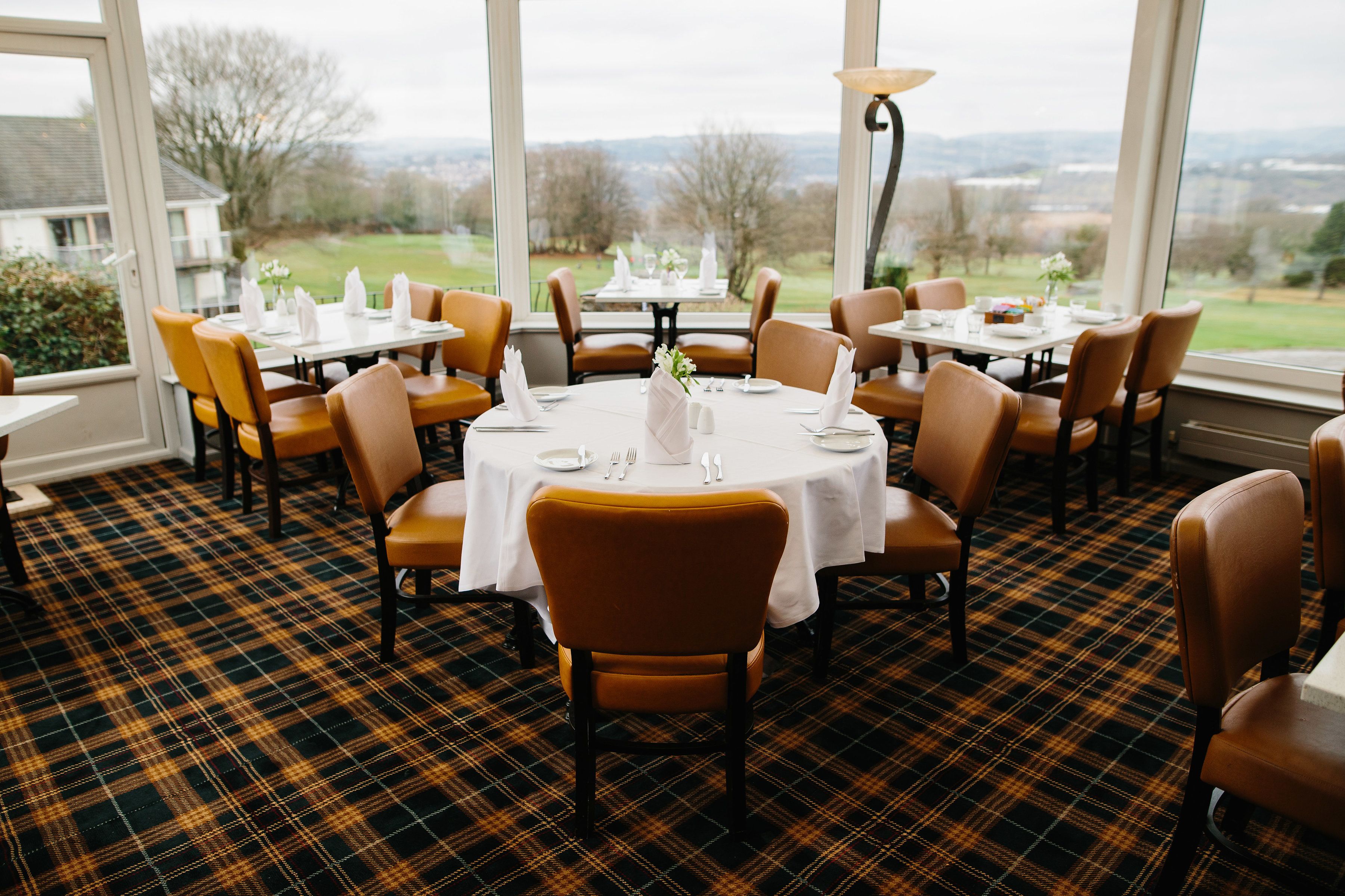 A restaurant with round tables and brown chairs, a plaid carpet, and a view of the countryside through large windows.