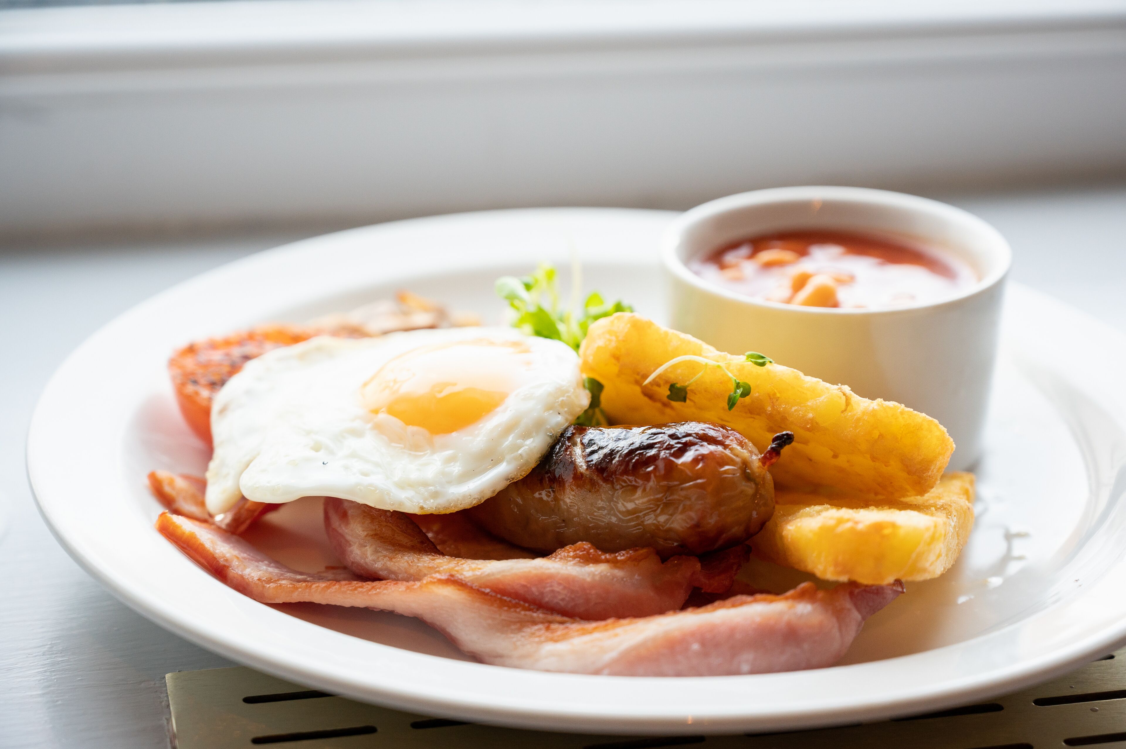 A plate of breakfast food including eggs, bacon, sausages, and fried plantains with a side of tomato sauce.