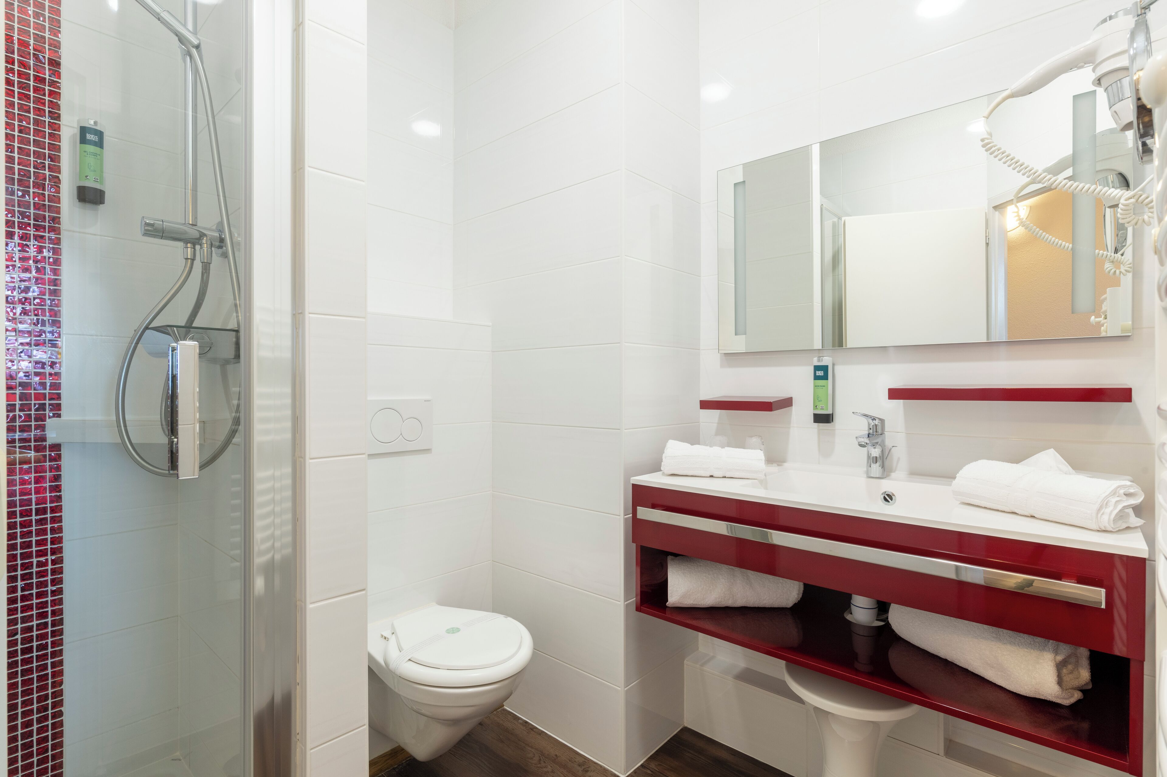 Et moderne badeværelse med en brusekabine i glas, et hvidt toilet og en vaskebordplads med rødt bord og hvid vask.