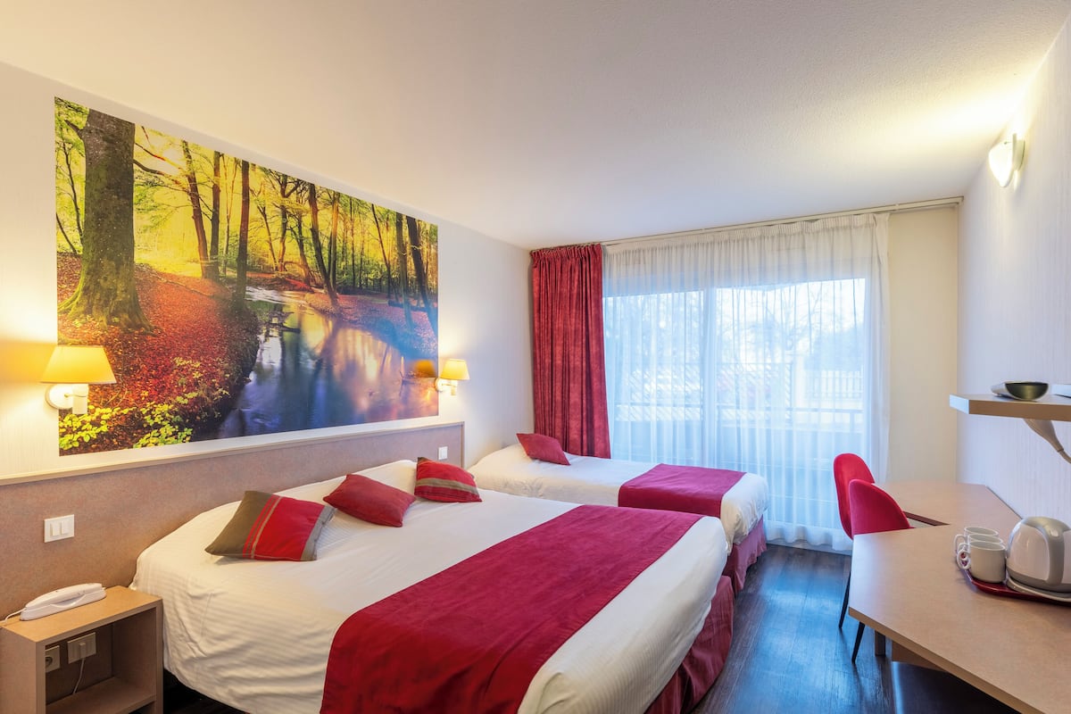 A hotel room with two beds, a desk, a chair, a painting of a forest, and a window with curtains.