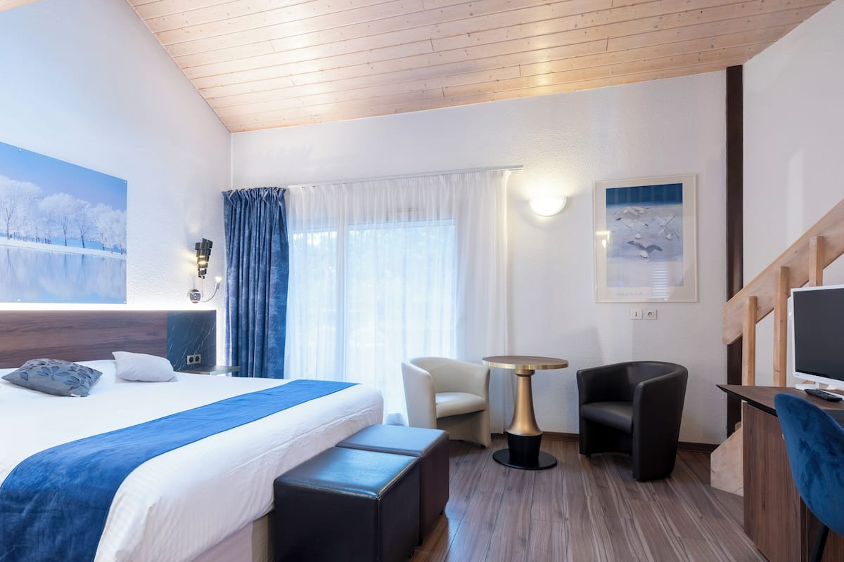A modern hotel room with a large bed, a desk, a chair, a small table, and a TV.
