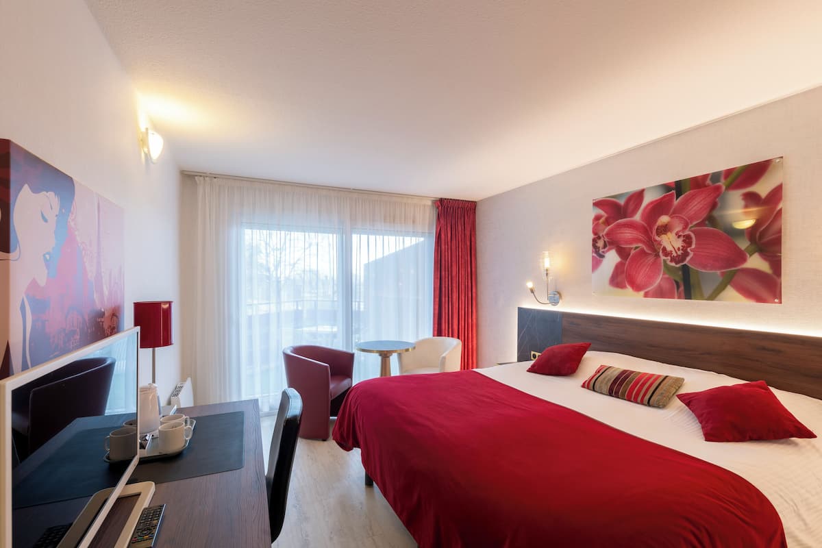 A hotel room with a bed, a desk with a laptop, a chair, a small table, and a window with curtains.