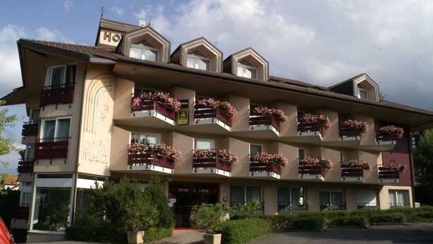 A multi-story hotel with balconies adorned with flower boxes, a visible entrance, and a clear sky. - Logis Hôtel Arc en Ciel (Thonon-les-Bains)